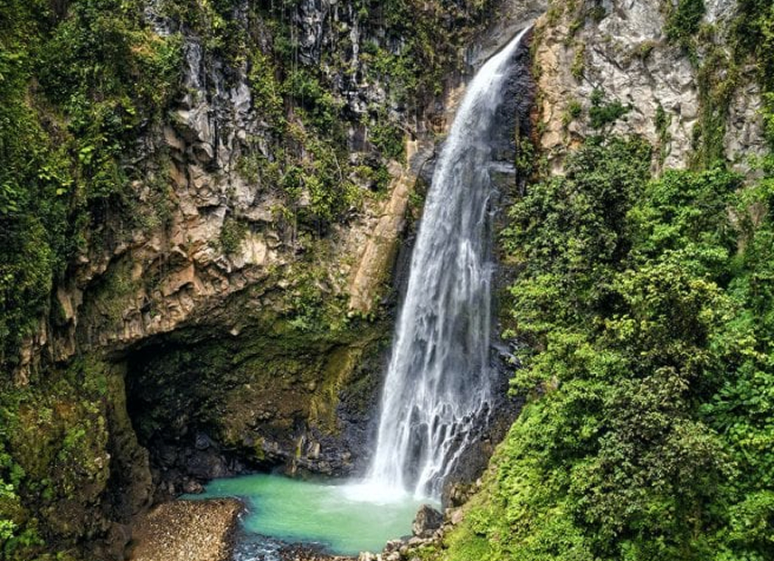 Tall waterfall flowing from a cliff into a turquoise pool surrounded by lush greenery, with a rock grotto carved into the hillside.