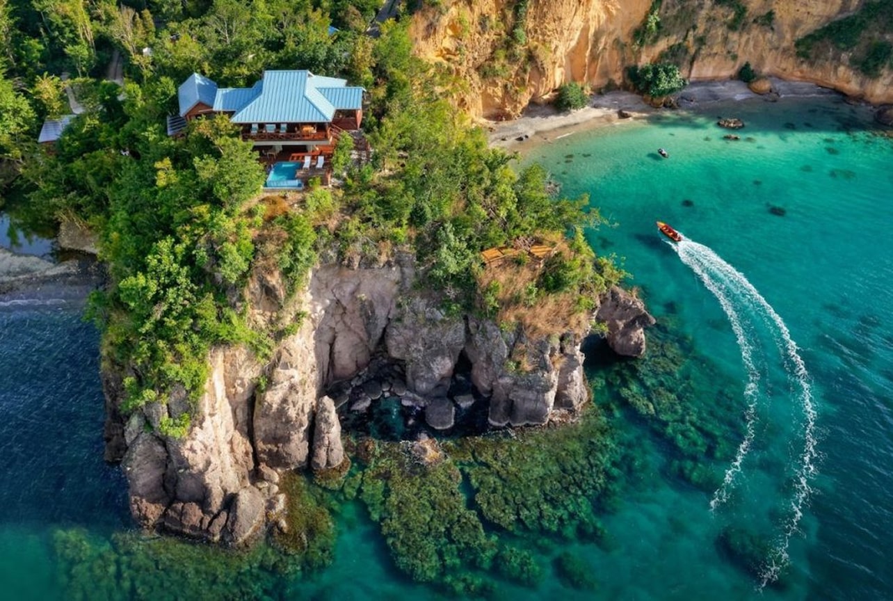 Villa built on a rocky cliff surrounded by forest, overlooking bright turquoise water, with a motorboat leaving a white wake across the bay.