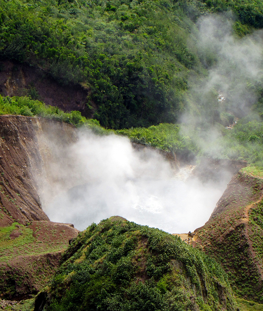 Steaming volcanic lake surrounded by steep green hillsides, with dense rainforest in the background.