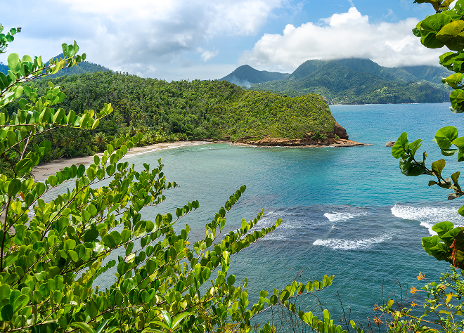 Scenic bay framed by green tropical hills and mountains, with turquoise waves breaking gently along the sandy shore.