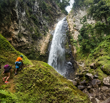 Two hikers climbing a mossy hillside near a tall, narrow waterfall cascading into a rocky pool below.