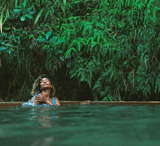 Woman relaxing in a natural hot spring pool, surrounded by dense green tropical plants and ferns.